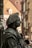 A detailed figure of a street performer, covered entirely in dark gray paint to resemble a statue, wearing historical attire with a hat and standing against an urban backdrop with blurred buildings.
