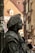A detailed figure of a street performer, covered entirely in dark gray paint to resemble a statue, wearing historical attire with a hat and standing against an urban backdrop with blurred buildings.