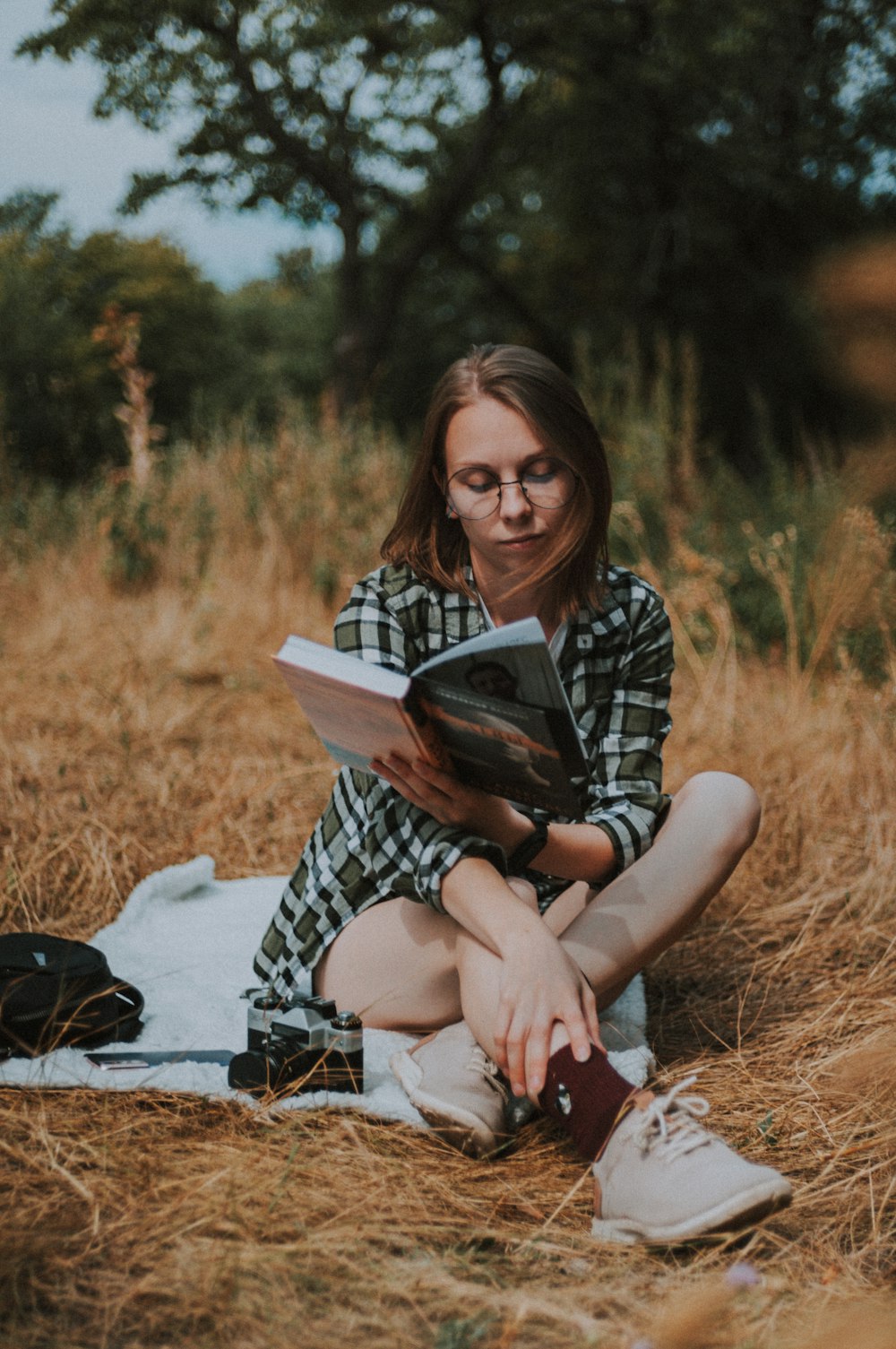 woman in black and white plaid dress shirt sitting on white textile reading book