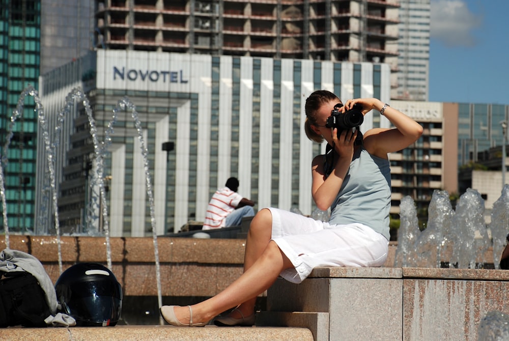 Frau in weißem Hemd und weißer Shorts sitzt auf Betonbank