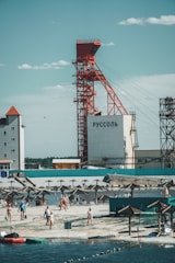 A beach scene with people enjoying recreational activities near the water. A large industrial structure with red framework stands prominently in the background, with buildings nearby. Sunshades line the beach, providing shade for the visitors.