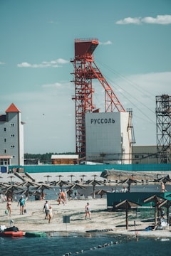 A beach scene with people enjoying recreational activities near the water. A large industrial structure with red framework stands prominently in the background, with buildings nearby. Sunshades line the beach, providing shade for the visitors.