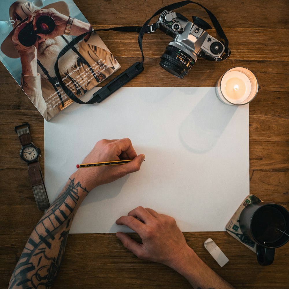 person holding white printer paper near black dslr camera and white ceramic mug