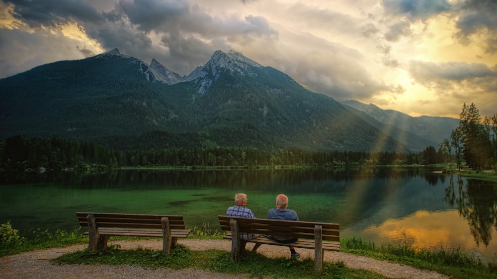 person sitting on brown wooden bench facing lake and mountains during daytime