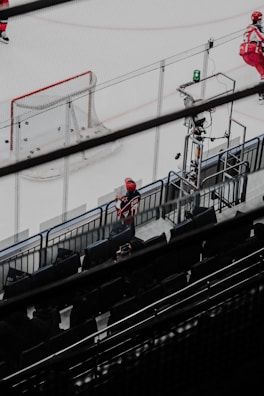A hockey rink seen from an elevated perspective, with a net on the left showing several pucks inside the goal. Players dressed in red uniforms are visible, some skating on the ice with sticks. The stands in the foreground are mostly empty, except for a few individuals, one wearing a red hat clearly visible.