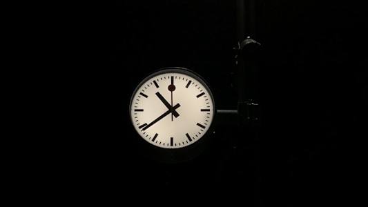 A round clock with a white face and black hour and minute marks is prominently displayed against a dark background. The clock has two black hands, indicating the time as approximately one minute past ten. A black support structure is attached to the right side of the clock.