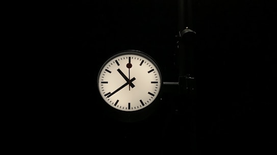 A round clock with a white face and black hour and minute marks is prominently displayed against a dark background. The clock has two black hands, indicating the time as approximately one minute past ten. A black support structure is attached to the right side of the clock.