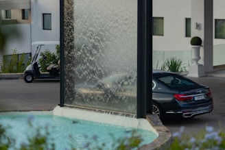 A modern water feature with a vertical glass panel where water cascades down into a pool. In the background, a black luxury sedan is parked beside a driveway, and a golf cart is visible near a building with white walls and windows. The area is landscaped with potted plants and greenery.