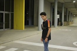 A young person stands in an urban environment with concrete and yellow structural elements visible. The individual is wearing a black T-shirt with colorful stripes on the sleeves and a small crossbody bag. The background includes large pillars, a glass door, and a pedestrian crossing on the ground.