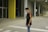 A young person stands in an urban environment with concrete and yellow structural elements visible. The individual is wearing a black T-shirt with colorful stripes on the sleeves and a small crossbody bag. The background includes large pillars, a glass door, and a pedestrian crossing on the ground.