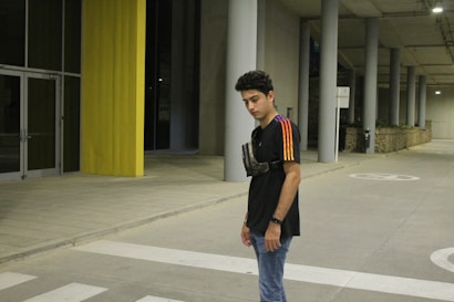A young person stands in an urban environment with concrete and yellow structural elements visible. The individual is wearing a black T-shirt with colorful stripes on the sleeves and a small crossbody bag. The background includes large pillars, a glass door, and a pedestrian crossing on the ground.