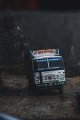 A large, colorful truck with various inscriptions and decorations is navigating a rough, dirt path on a foggy day. The words 'ROAD KING' are prominently displayed on the bumper, and the truck appears to be traveling in a mountainous region surrounded by dark, rocky terrain.