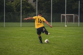 man in yellow and black soccer jersey kicking soccer ball on green grass field during daytime