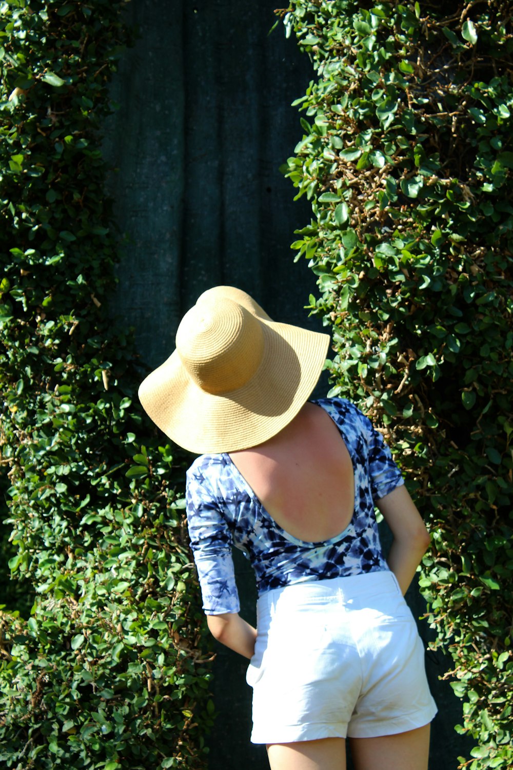 woman in white and blue floral dress wearing brown hat standing near green plant