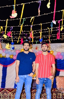 Two people are standing in a festive setting with colorful decorations. The background features hanging ribbons and lights, along with balloons in pastel colors. Both individuals are casually dressed in jeans and t-shirts.