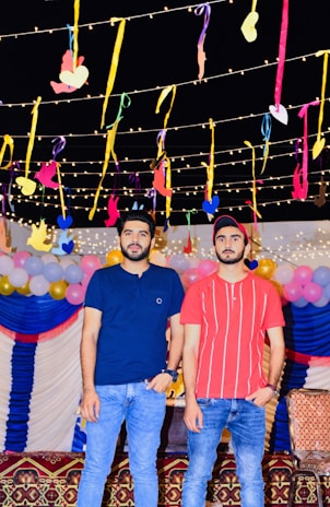 Two people are standing in a festive setting with colorful decorations. The background features hanging ribbons and lights, along with balloons in pastel colors. Both individuals are casually dressed in jeans and t-shirts.
