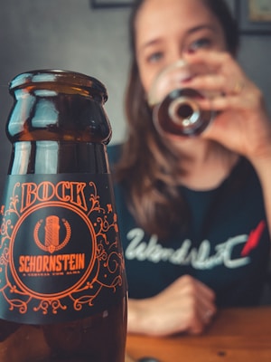A beer bottle with ornate orange lettering stands prominently in the foreground, with a person in the background drinking from a glass. The label reads 'Bock Schornstein.' The person is slightly out of focus, wearing a dark shirt with a white design.