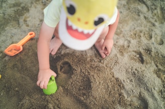 A child wearing a dinosaur hat is playing in the sand. The child is using a green sand mold and there is an orange shovel nearby. The sand appears to be in a sandbox or at a beach.