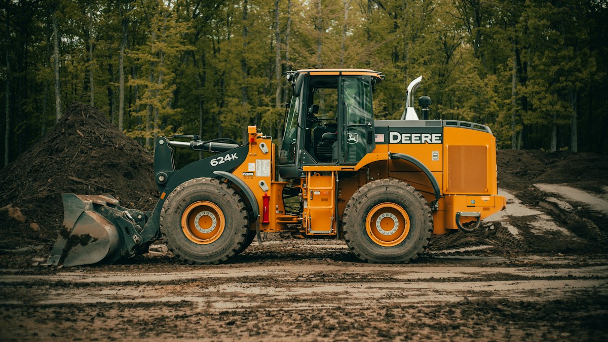 Wheel loader with its bucket lowered beside soil stockpiles and trees.