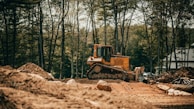 A yellow bulldozer is situated in a clearing surrounded by tall trees, pushing large piles of dirt and rocks at a construction site. The setting is outdoors with a forested background, and the atmosphere seems industrious as the machine is actively working on the earth.