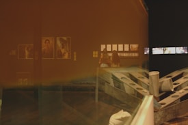 The image captures a dimly lit interior space, possibly an art gallery or museum, featuring two people observing framed photographs and exhibits on the walls. There is a reflection on the glass surface, creating a layered effect. Artworks and installation pieces are prominent, set against a dark brown or orange-toned background.