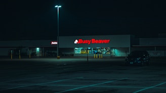 A dimly lit parking lot at night with a store named 'Busy Beaver' illuminated in the background. The building is surrounded by a few parked cars and pole lights that cast a soft glow.