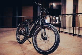A rugged mountain bike with thick tires is parked in an urban setting, with a metal railing in the background. The scene is dimly lit, suggesting it was taken during the evening or at night, highlighting the bike's robust construction and reflective elements.