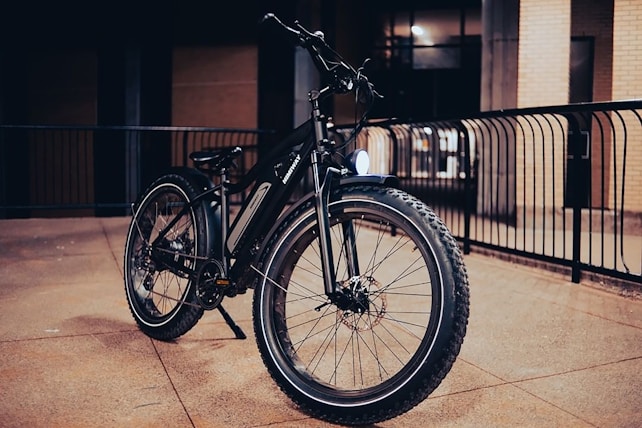 A rugged mountain bike with thick tires is parked in an urban setting, with a metal railing in the background. The scene is dimly lit, suggesting it was taken during the evening or at night, highlighting the bike's robust construction and reflective elements.