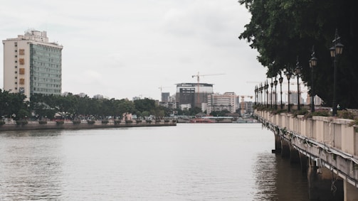 A waterfront view features a large body of calm water bordered by a railing with lampposts. There is a prominent high-rise building on the left, surrounded by trees and other urban structures. The sky is overcast, with cranes visible in the distance, suggesting ongoing construction.