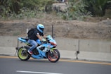 A motorcyclist is riding a brightly colored sport bike along a road bordered by a concrete barrier and dense foliage. The rider is wearing a light blue helmet, black jacket, jeans, and red shoes. The motorcycle is predominantly blue with accents of green and red, featuring a sleek design.