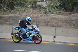 A motorcyclist is riding a brightly colored sport bike along a road bordered by a concrete barrier and dense foliage. The rider is wearing a light blue helmet, black jacket, jeans, and red shoes. The motorcycle is predominantly blue with accents of green and red, featuring a sleek design.