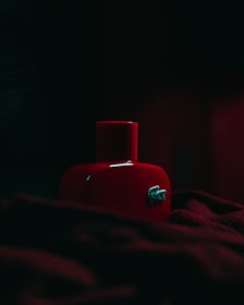 A red bottle with a glossy finish is partially illuminated in a dimly lit setting. The bottle features a crocodile logo and is surrounded by soft, dark fabric.