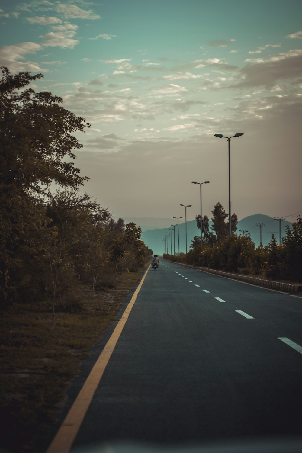 gray concrete road between green trees under white clouds and blue sky during daytime