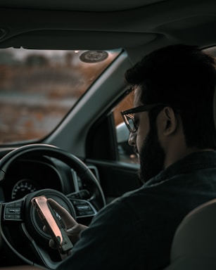 A man is sitting in the driver's seat of a vehicle, focusing intently on his smartphone. He is wearing glasses and has a beard, with a relaxed posture. The interior of the vehicle is visible, showing the steering wheel and dashboard.