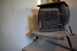 A vintage black cast iron boxwood stove with the brand name and model number embossed on its front is placed against a plain light-colored wall. The stove has a circular vent on its door and sturdy, simple legs supporting its body. There is a subtle shadow cast on the wall.