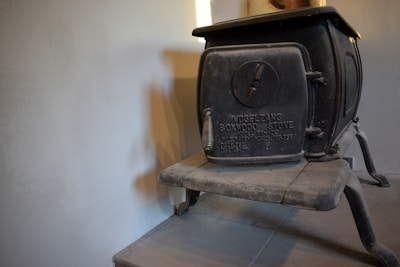 A vintage black cast iron boxwood stove with the brand name and model number embossed on its front is placed against a plain light-colored wall. The stove has a circular vent on its door and sturdy, simple legs supporting its body. There is a subtle shadow cast on the wall.