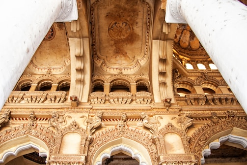 Ornate architectural details featuring grand columns, intricately carved arches, and decorative sculptures on a historical building ceiling. The design includes elaborate carvings and patterns, showcasing artistic craftsmanship.