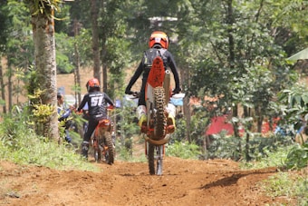A group of dirt bikers navigate a forest trail, with one rider performing a wheelie. The scene is vibrant with green foliage and trees surrounding the path. The riders are dressed in protective gear and helmets, and the trail is a reddish-brown dirt path.
