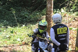 Two individuals in motocross gear are in a forested area. One person is seated on a dirt bike while the other stands nearby, wearing matching gear. Bright, leafy greens of the forest and clothing logos are visible.