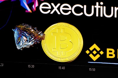 A shiny gold coin featuring the Bitcoin logo is placed next to a colorful, geometric crystal. The background includes branding elements with the Binance logo and the word 'executium'. A faint chart is visible in the background, indicating a financial or trading context.