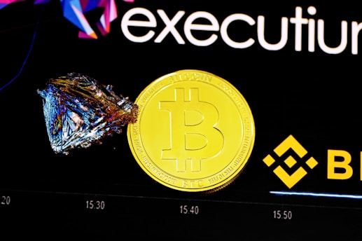 A shiny gold coin featuring the Bitcoin logo is placed next to a colorful, geometric crystal. The background includes branding elements with the Binance logo and the word 'executium'. A faint chart is visible in the background, indicating a financial or trading context.