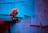 A small robot figure sits at the edge of a table, surrounded by various notes and papers pinned to the wall. The papers contain motivational phrases and doodles. The setting is dimly lit with a combination of blue and red lighting, creating a moody atmosphere.