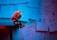A small robot figure sits at the edge of a table, surrounded by various notes and papers pinned to the wall. The papers contain motivational phrases and doodles. The setting is dimly lit with a combination of blue and red lighting, creating a moody atmosphere.
