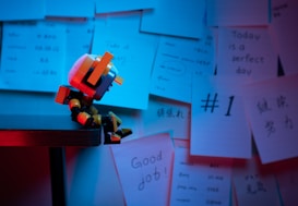 A small robot figure sits at the edge of a table, surrounded by various notes and papers pinned to the wall. The papers contain motivational phrases and doodles. The setting is dimly lit with a combination of blue and red lighting, creating a moody atmosphere.