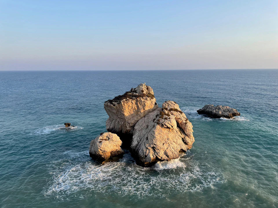 Large rocky outcropping juts into ocean, surrounded by three varying-sized boulders and clear blue sky