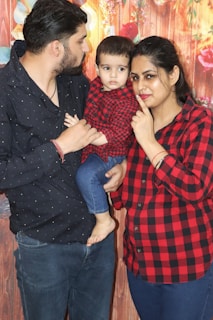 A family poses together with a man holding a young child dressed in red and black checkered clothing, while a woman stands beside them, also wearing a similar patterned shirt. The background is decorated with flowers and wooden textures.