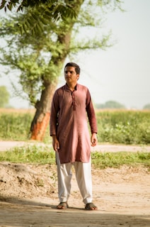 A person stands outdoors on a dirt path, with lush green fields and trees in the background. They are wearing a maroon kurta and white pants, and appear to be gazing into the distance.