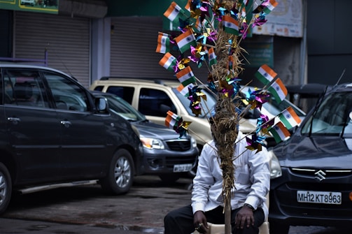 A street scene featuring a person sitting on a chair partially hidden by a large decorative branch adorned with Indian flags and colorful pinwheels. The area appears to be a market or urban setting with several parked cars and closed shop shutters in the background.