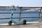 A teal electric scooter is parked on a paved walkway next to a metal railing overlooking a large body of water. In the background, there's a boat on the water and a cityscape with numerous buildings under a clear blue sky.