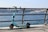 A teal electric scooter is parked on a paved walkway next to a metal railing overlooking a large body of water. In the background, there's a boat on the water and a cityscape with numerous buildings under a clear blue sky.